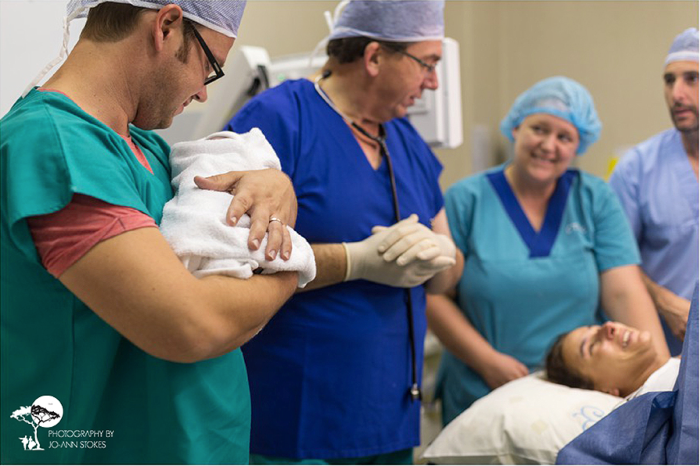 A father in theatre scrubs holds his newborn while the doctor, nurse and mother look on